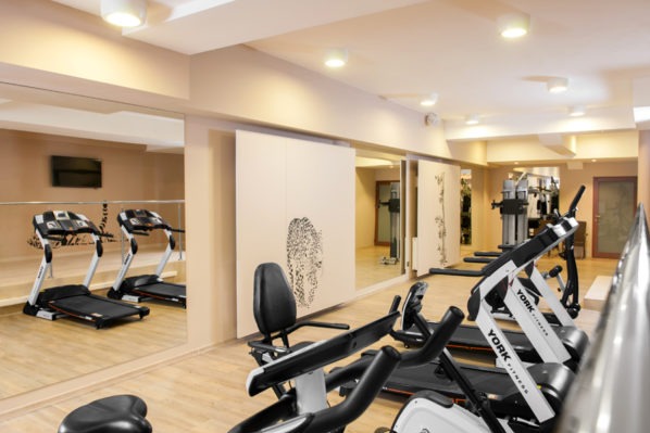 Jasne wnętrze siłowni z bieżniami York Fitness, rowerami stacjonarnymi i lustrami, w których odbijają się kolejne urządzenia. Na ścianie dekoracyjne panele z motywem graficznym.