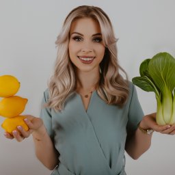 RÓWNOWAGA Julia Bednarkiewicz - Uśmiechnięta blondynka w turkusowym stroju prezentuje w dłoniach piramidę z trzech cytryn i pak choi na białym tle.