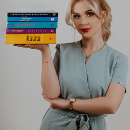 RÓWNOWAGA Julia Bednarkiewicz - Uśmiechnięta blondynka w turkusowym uniformie trzymająca stos książek o tematyce dietetycznej, z widocznym złotym zegarkiem i bransoletką