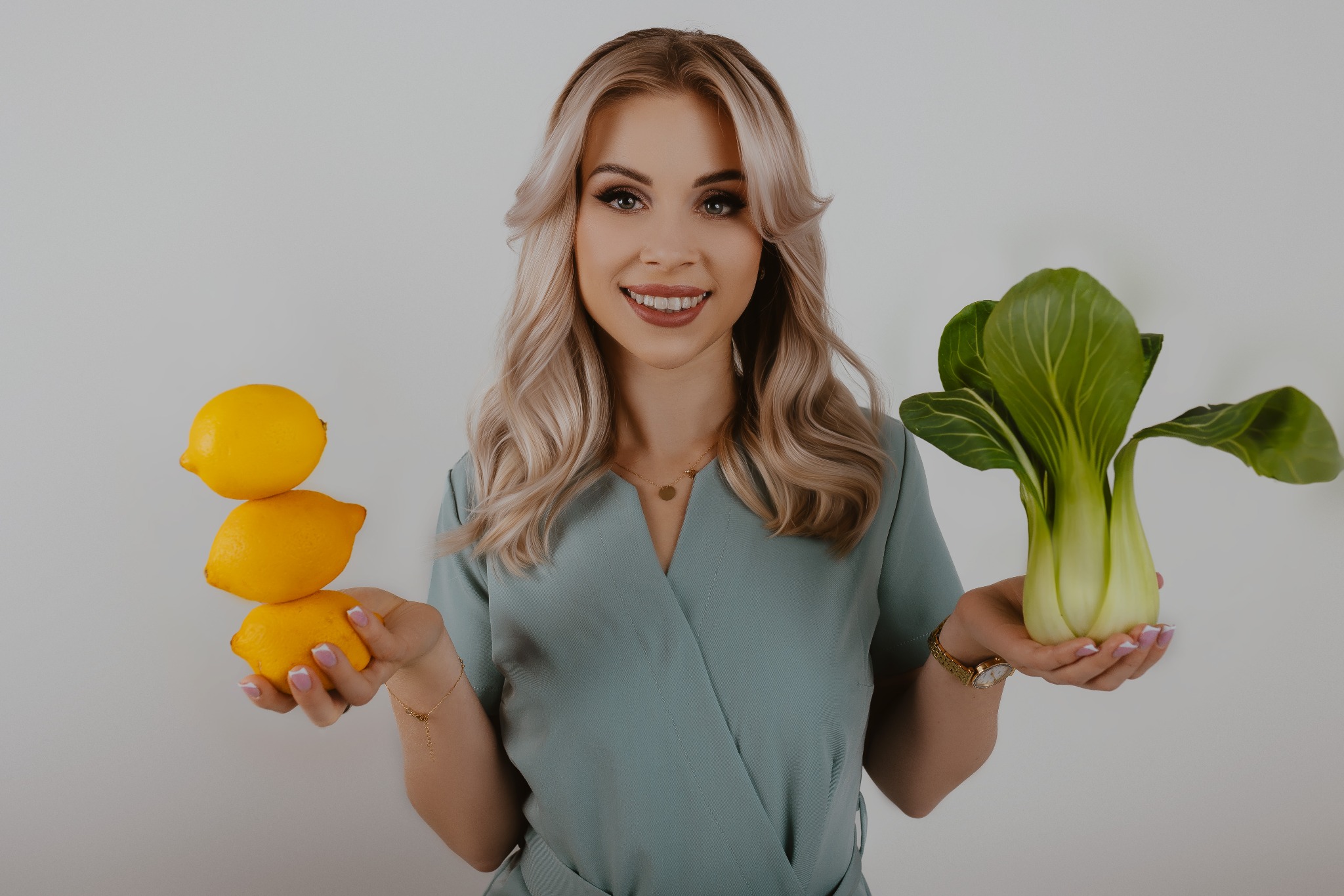 Uśmiechnięta blondynka w turkusowym stroju prezentuje w dłoniach piramidę z trzech cytryn i pak choi na białym tle.