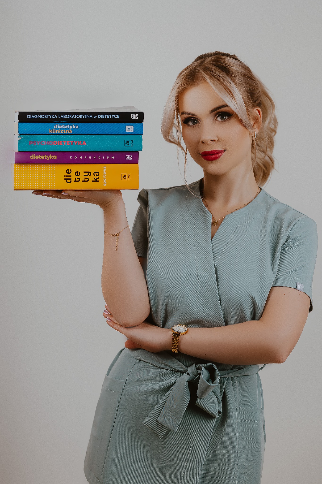 Uśmiechnięta blondynka w turkusowym uniformie trzymająca stos książek o tematyce dietetycznej, z widocznym złotym zegarkiem i bransoletką