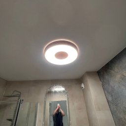 Daneks - Nowoczesna łazienka z okrągłą lampą sufitową i owalnym lustrem w folii ochronnej, odbijającym fotografa. Widoczna kabina prysznicowa i szara ściana z płytek.