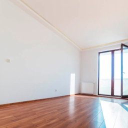 Daneks - Jasne, puste wnętrze z drewnianą podłogą i białymi ścianami. Widoczne grzejniki, otwarte drzwi balkonowe i listwy przypodłogowe w kolorze drewna. Świeże, minimalistyczne wykończenie.