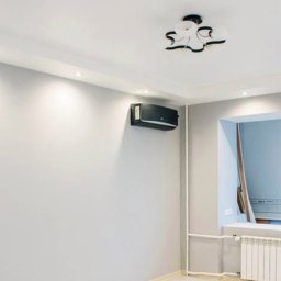 Daneks - Jasne, odświeżone wnętrze pokoju z szarą ścianą z wzorem, klimatyzacją i grzejnikiem. Widoczne oświetlenie punktowe i nowoczesna lampa sufitowa.