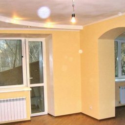 Daneks - Jasne wnętrze po remoncie w Gdańsku: żółte ściany, białe okna z grzejnikami. Widoczny łuk między pokojami i podwieszany sufit z oświetleniem.