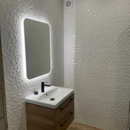 Nowoczesna łazienka z umywalką na szafce z drewnopodobnym frontem, podświetlanym lustrem i trójwymiarowymi białymi płytkami na ścianach.