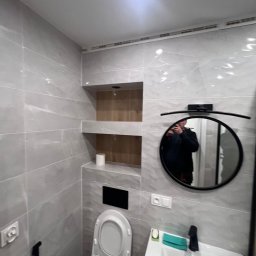 Daneks - Wykończona łazienka w szarościach z toaletą, umywalką i okrągłym lustrem. W ścianie wnęka z półkami wyłożona drewnem. Widoczne gniazdka elektryczne.