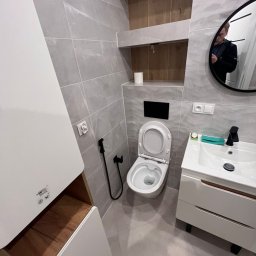 Daneks - Jasna łazienka po remoncie: szare płytki, biała umywalka z szafką, toaleta z bidetką, lustro z odbiciem osoby i drewniana wnęka z półkami.