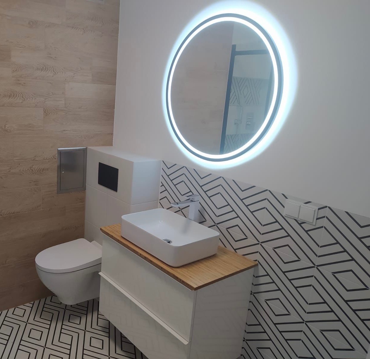 Nowoczesna łazienka z umywalką nablatową na drewnianym blacie, okrągłym lustrem z podświetleniem LED i geometrycznymi płytkami na podłodze i ścianie.