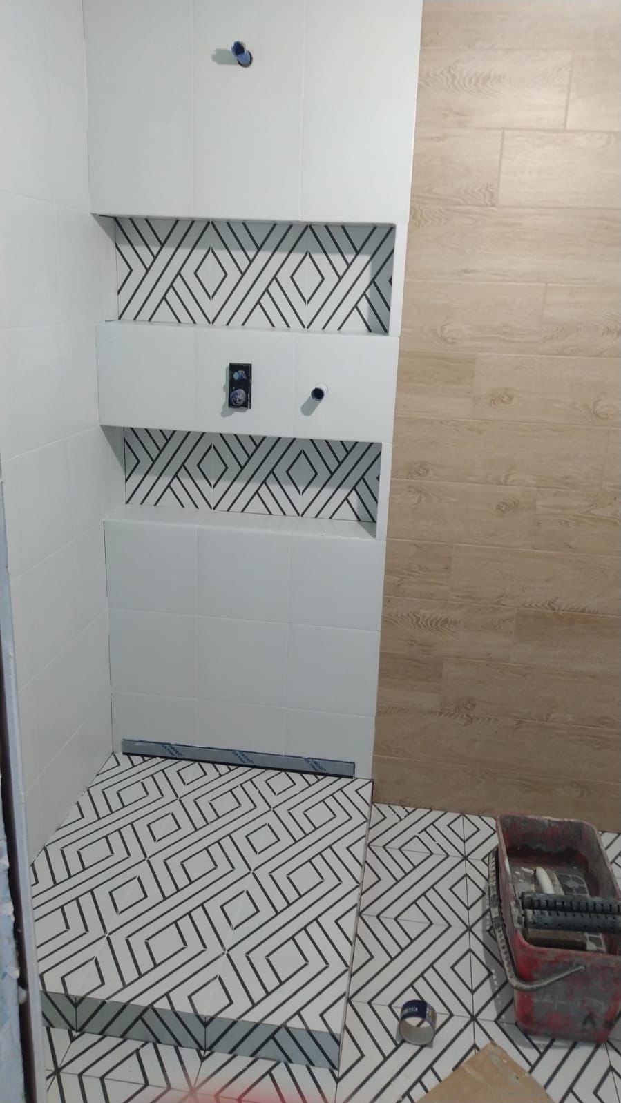 Wnętrze łazienki w trakcie wykańczania: białe płytki z wzorem na ścianach, drewnopodobne panele, wzorzysta podłoga. Widoczny pojemnik z narzędziami.