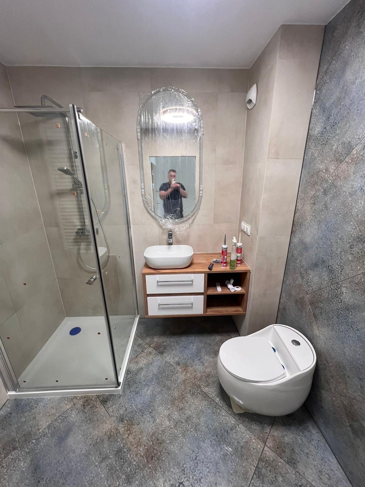 Wykończona łazienka z kabiną prysznicową, umywalką na szafce z drewnianym blatem i toaletą. Lustro owinięte folią ochronną. Dekoracyjne płytki podłogowe i ścienne.