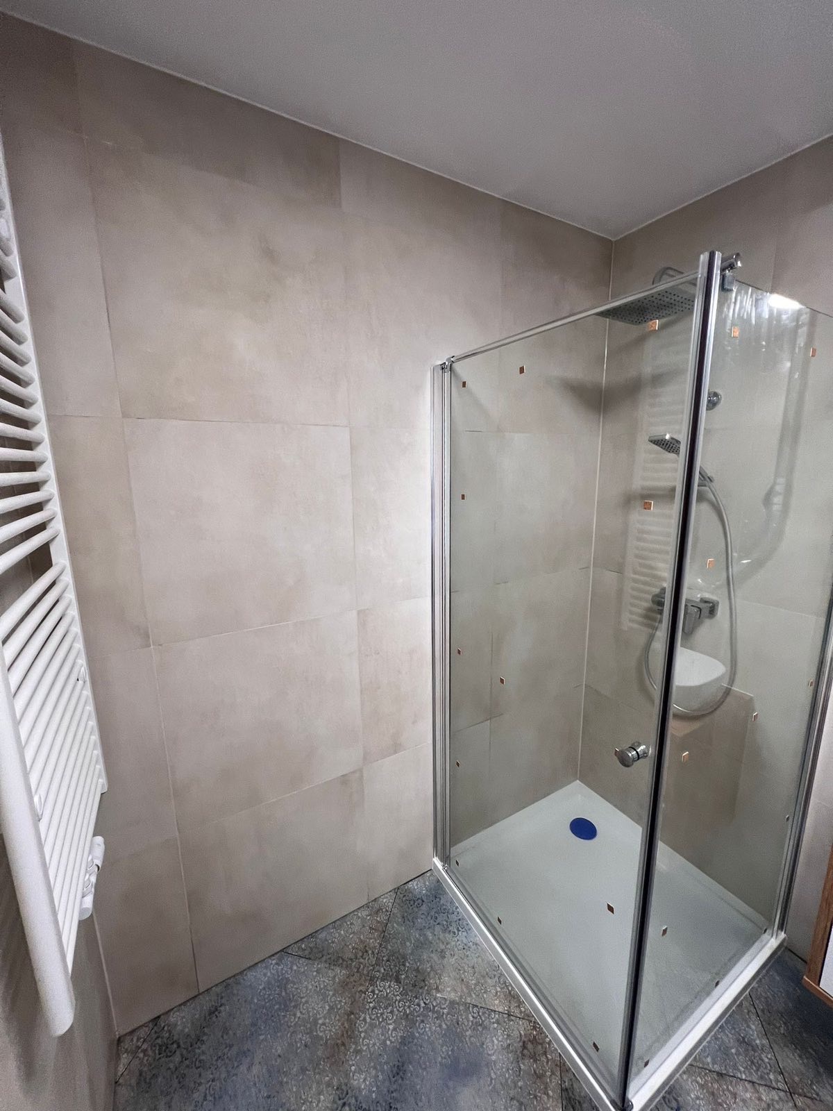 Jasna łazienka z kabiną prysznicową ze szklanymi drzwiami, grzejnikiem drabinkowym i płytkami imitującymi kamień na ścianach oraz wzorzystą podłogą.