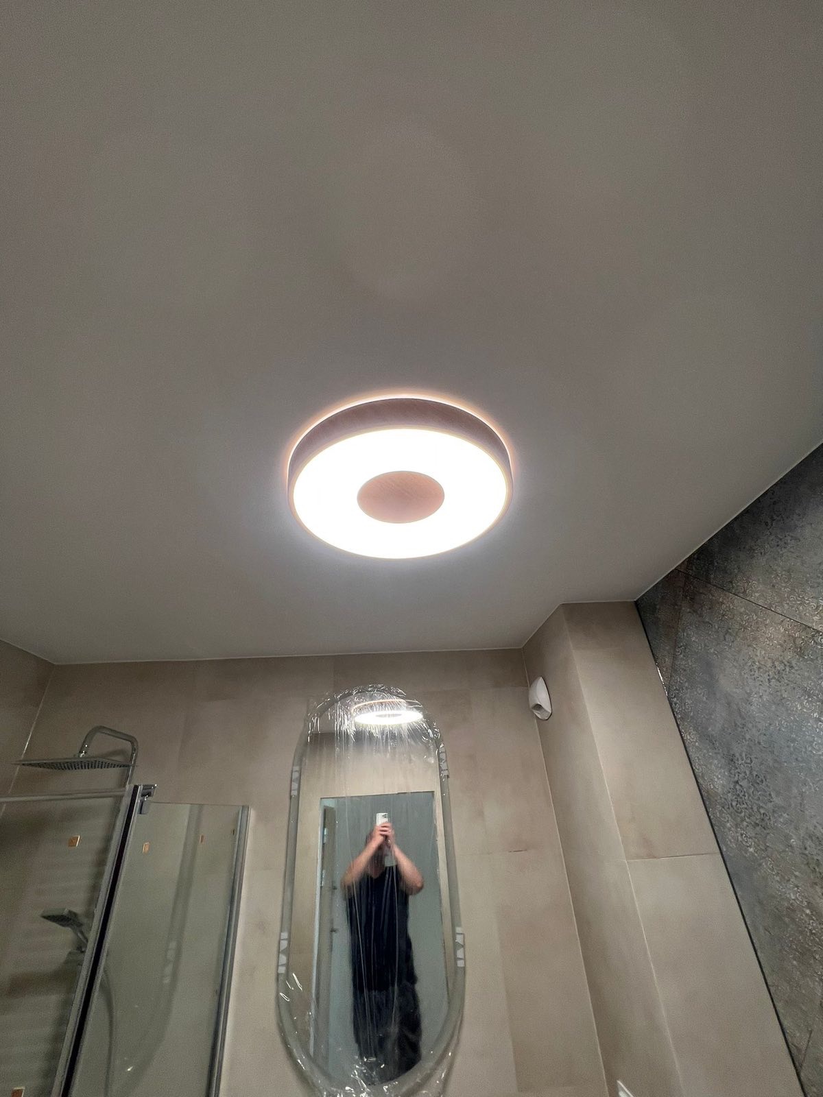 Nowoczesna łazienka z okrągłą lampą sufitową i owalnym lustrem w folii ochronnej, odbijającym fotografa. Widoczna kabina prysznicowa i szara ściana z płytek.