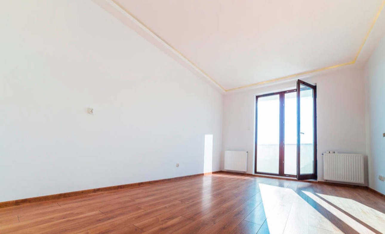 Jasne, puste wnętrze z drewnianą podłogą i białymi ścianami. Widoczne grzejniki, otwarte drzwi balkonowe i listwy przypodłogowe w kolorze drewna. Świeże, minimalistyczne wykończenie.