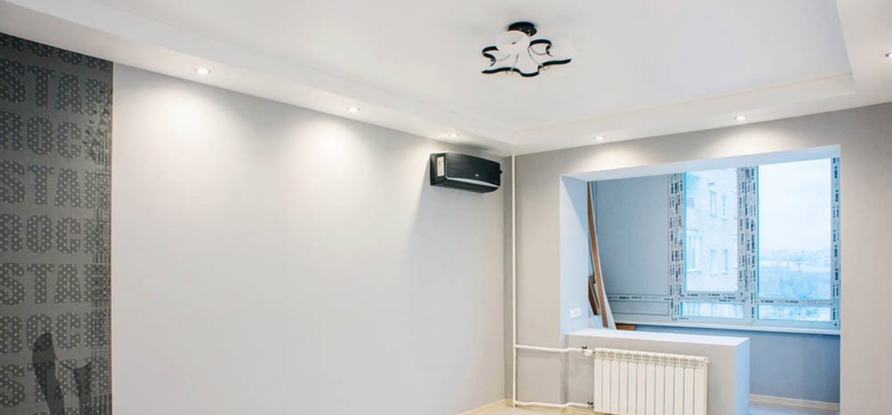 Jasne, odświeżone wnętrze pokoju z szarą ścianą z wzorem, klimatyzacją i grzejnikiem. Widoczne oświetlenie punktowe i nowoczesna lampa sufitowa.