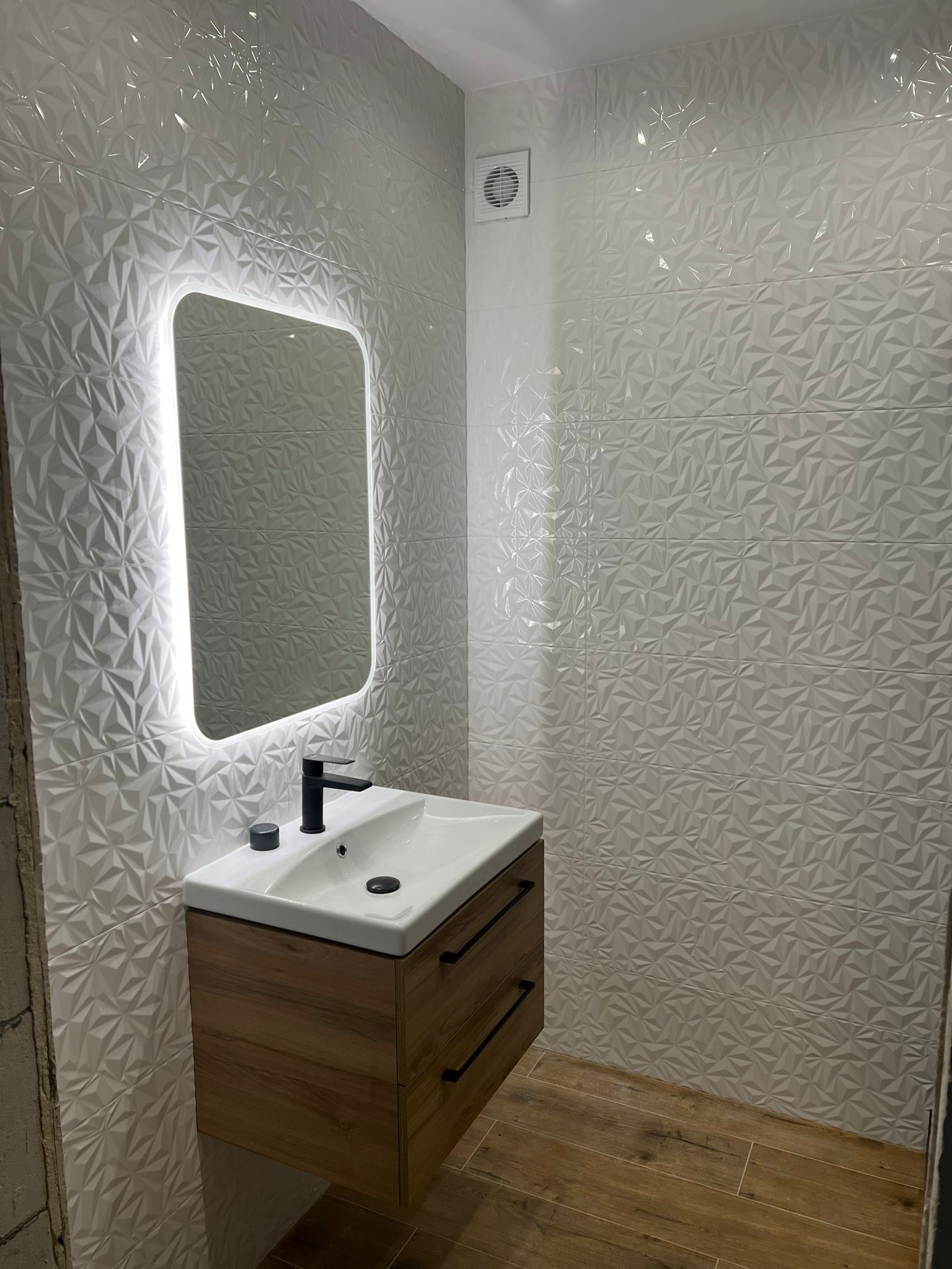 Nowoczesna łazienka z umywalką na szafce z drewnopodobnym frontem, podświetlanym lustrem i trójwymiarowymi białymi płytkami na ścianach.