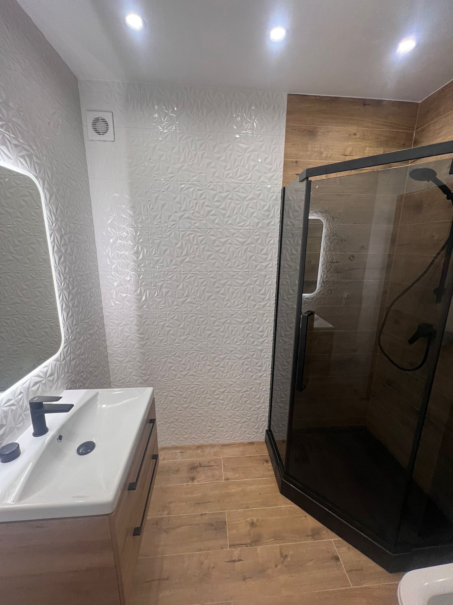 Nowoczesna łazienka po remoncie: kabina prysznicowa z czarnymi ramami, umywalka z szafką w odcieniach drewna, białe płytki 3D na ścianie i drewnopodobna podłoga.