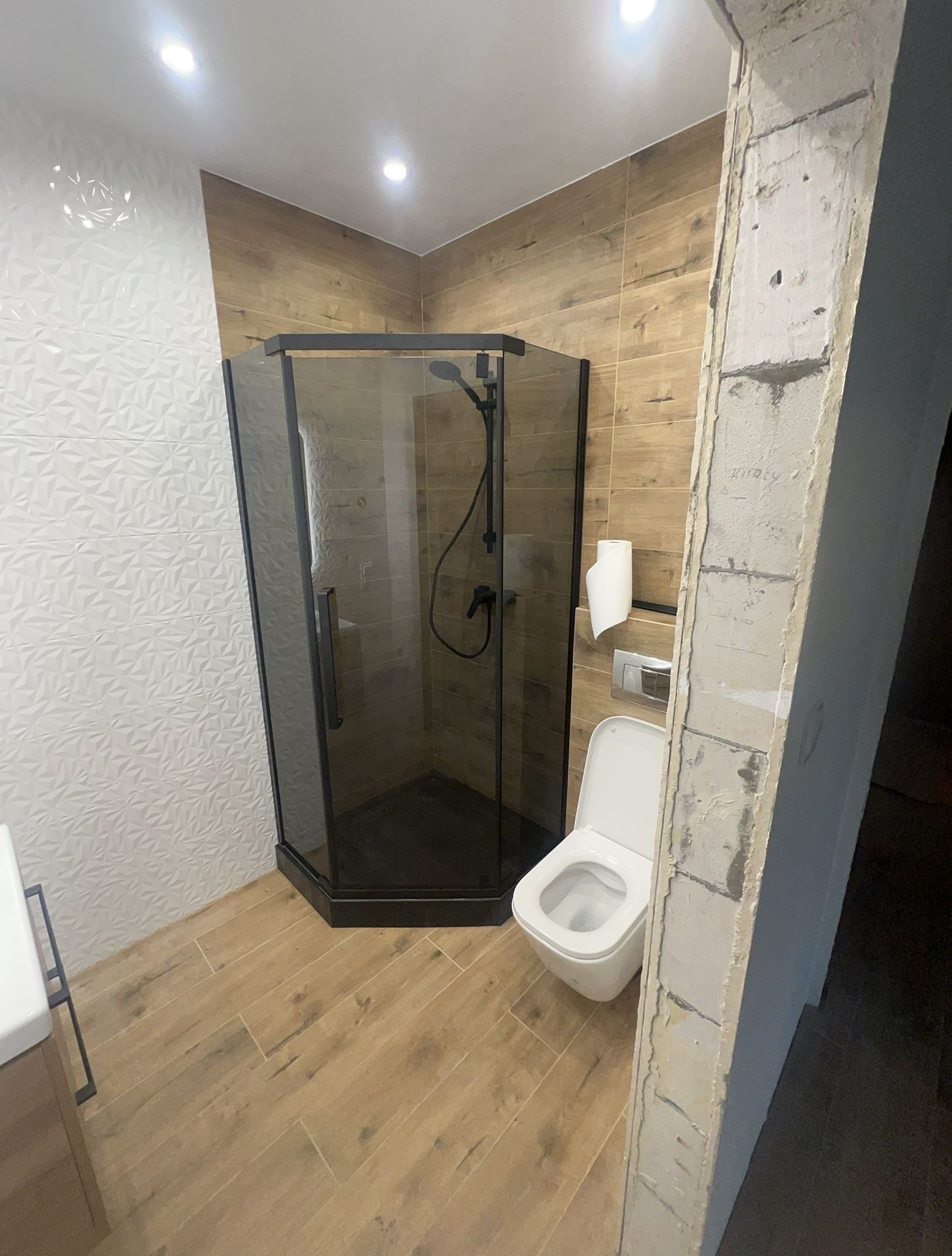 Nowoczesna łazienka po remoncie: kabina prysznicowa z czarnymi profilami, toaleta, drewnopodobne płytki i dekoracyjna ściana z białymi trójwymiarowymi płytkami.