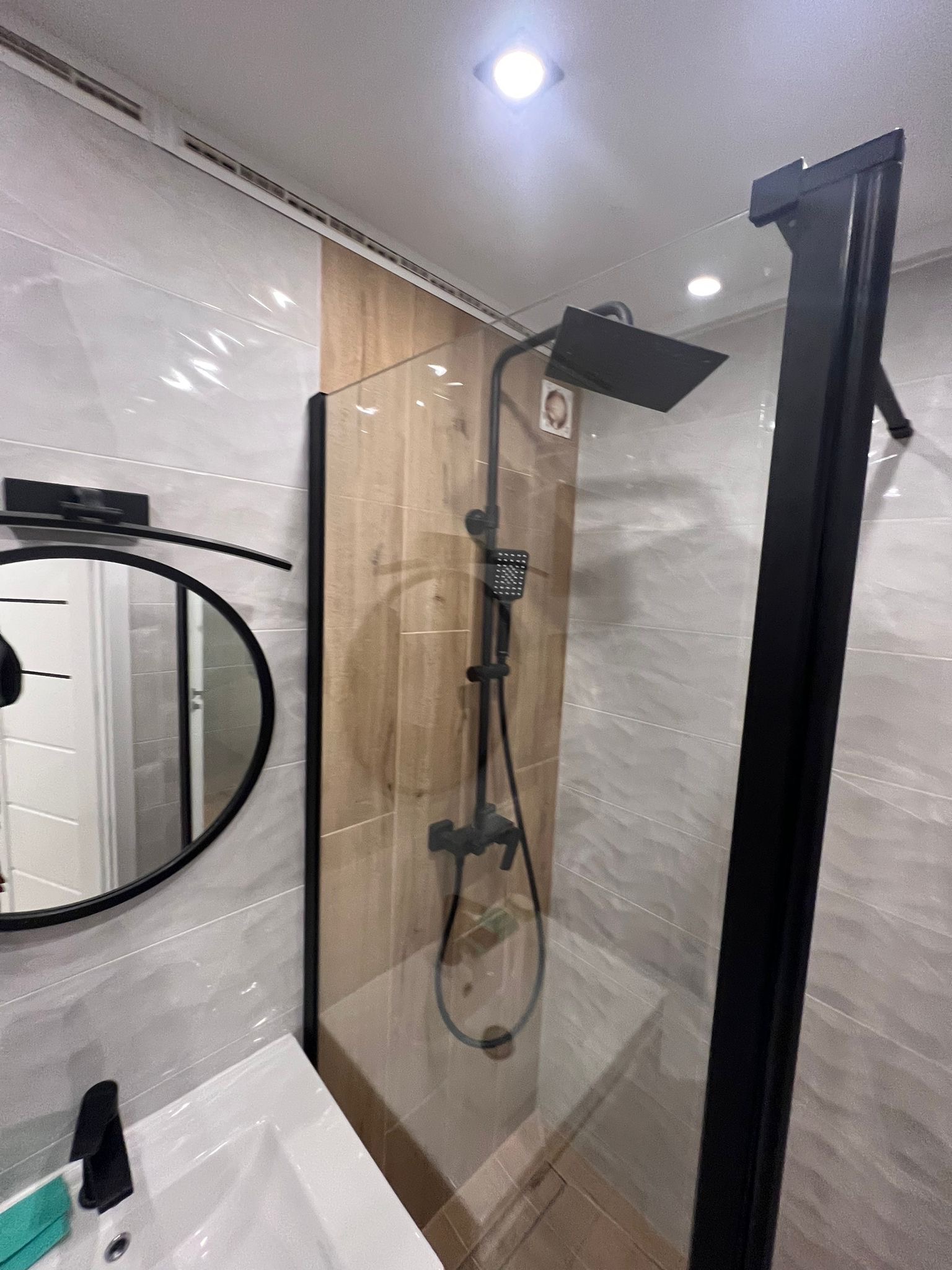 Nowoczesna łazienka po remoncie: kabina prysznicowa typu walk-in z czarną armaturą, lustro w czarnej ramie, umywalka, płytki imitujące drewno i fale, oświetlenie punktowe.