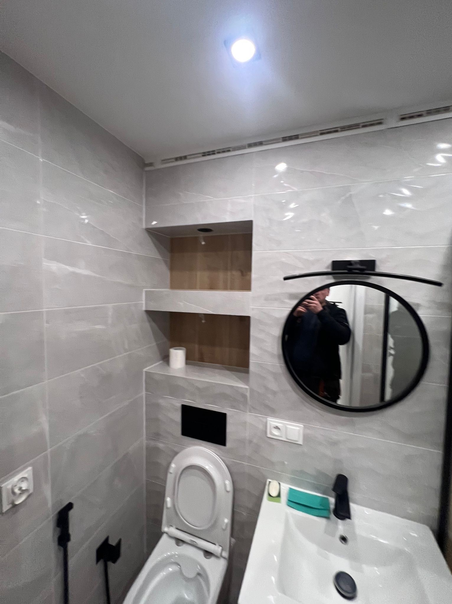 Wykończona łazienka w szarościach z toaletą, umywalką i okrągłym lustrem. W ścianie wnęka z półkami wyłożona drewnem. Widoczne gniazdka elektryczne.