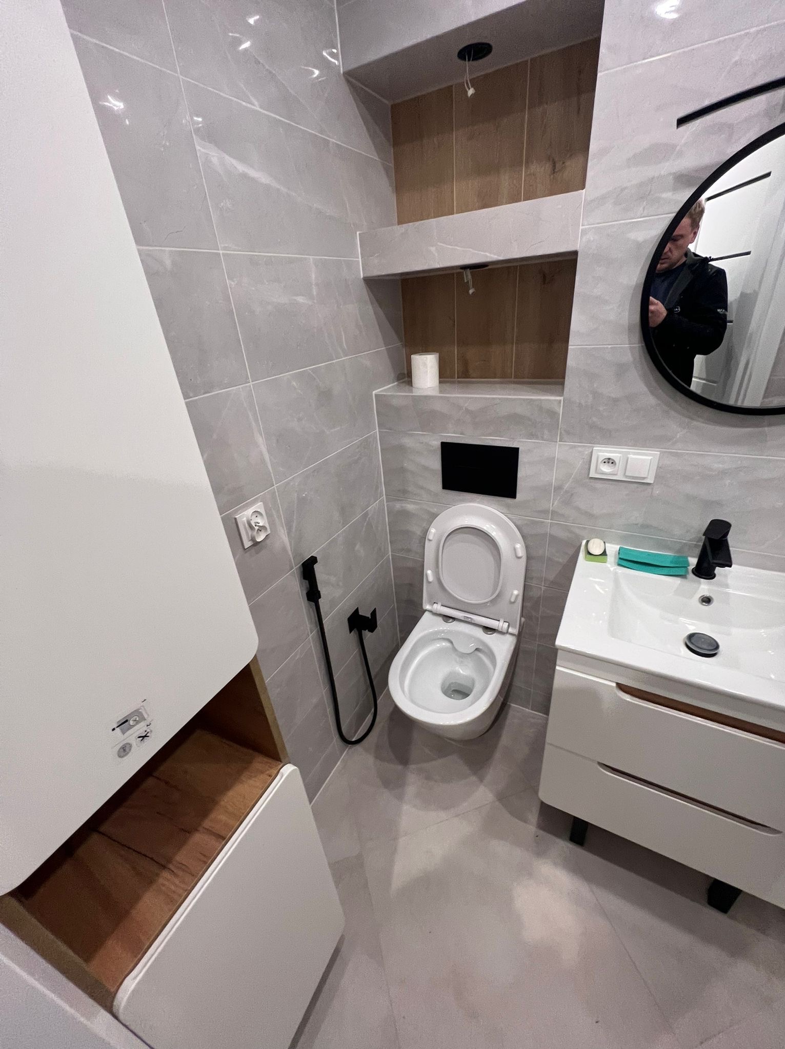 Jasna łazienka po remoncie: szare płytki, biała umywalka z szafką, toaleta z bidetką, lustro z odbiciem osoby i drewniana wnęka z półkami.
