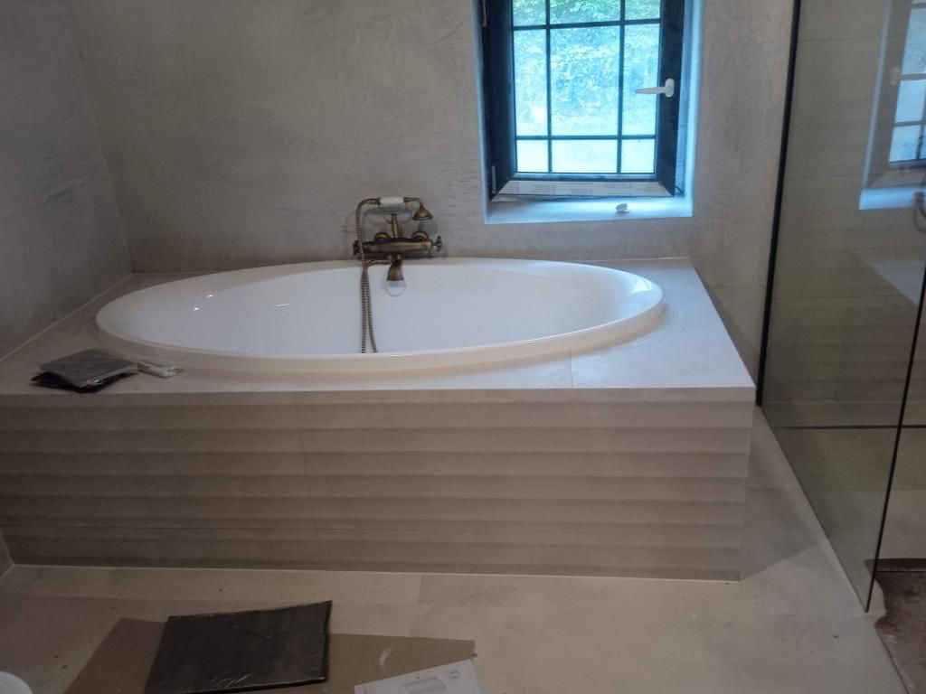 Wykończona owalna wanna z baterią w stylu retro, obudowana płytkami z poziomymi żłobieniami. Obok okno, kabina prysznicowa i podłoga w jasnym odcieniu beżu.