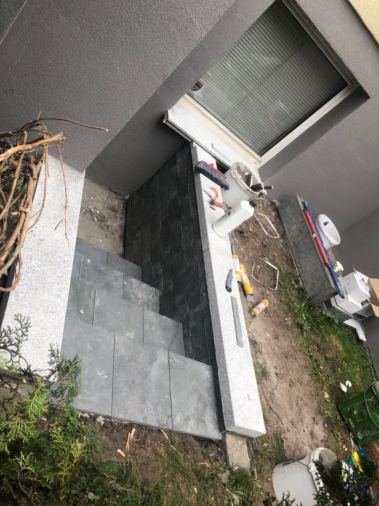 Wykończenie schodów zewnętrznych z kamienia. Widoczne narzędzia i materiały budowlane. Schody prowadzą do drzwi z roletą. Ujęcie z góry.