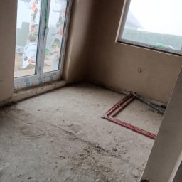 Tynk-daw - Surowe wnętrze pokoju w trakcie prac wykończeniowych, ściany pokryte świeżym tynkiem, widoczne okna PCV, instalacja grzewcza w podłodze i surowe drewniane belki stropowe.