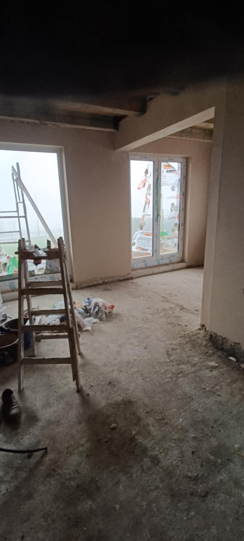 Wnętrze w trakcie prac wykończeniowych: ściany pokryte jasnym tynkiem, drewniana drabina oparta o ścianę przy oknie, widoczne wiadra i folie ochronne.