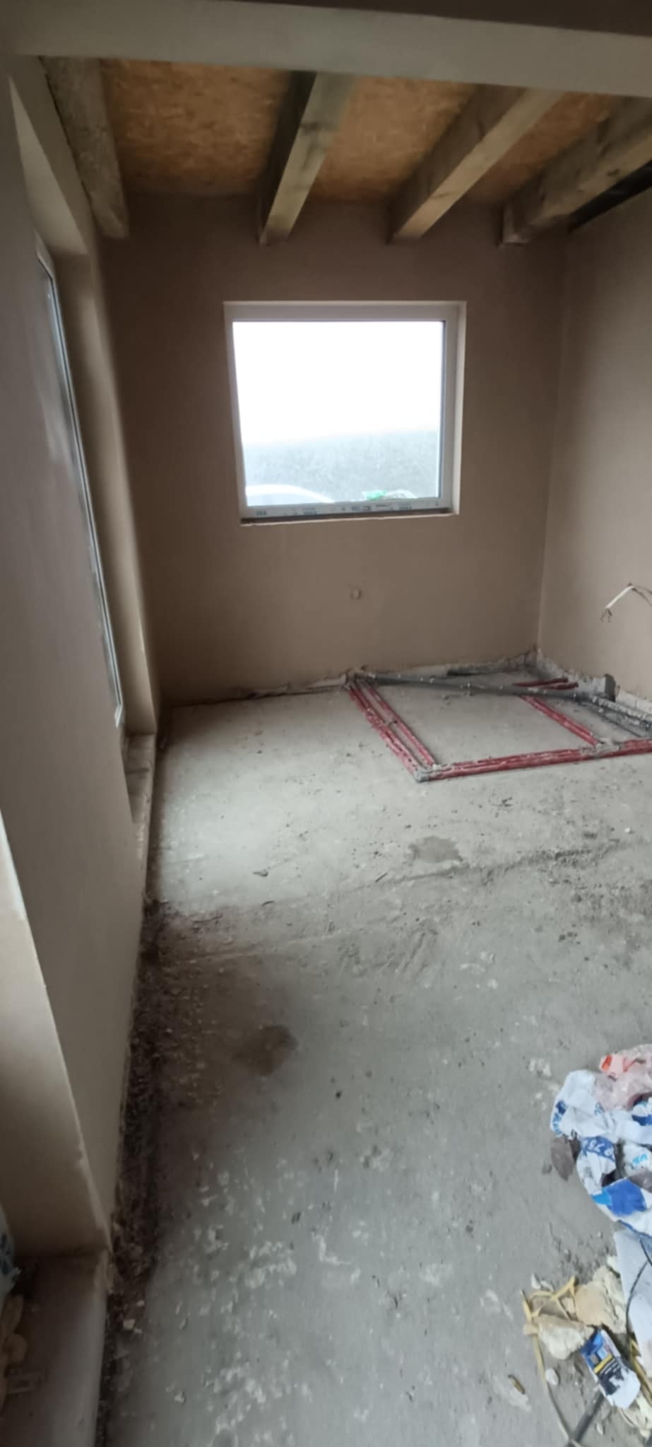 Surowe wnętrze pokoju po tynkowaniu maszynowym: ściany pokryte świeżym tynkiem, widoczne instalacje grzewcze podłogowe, sufit z drewnianych belek, okno z widokiem na zewnątrz.