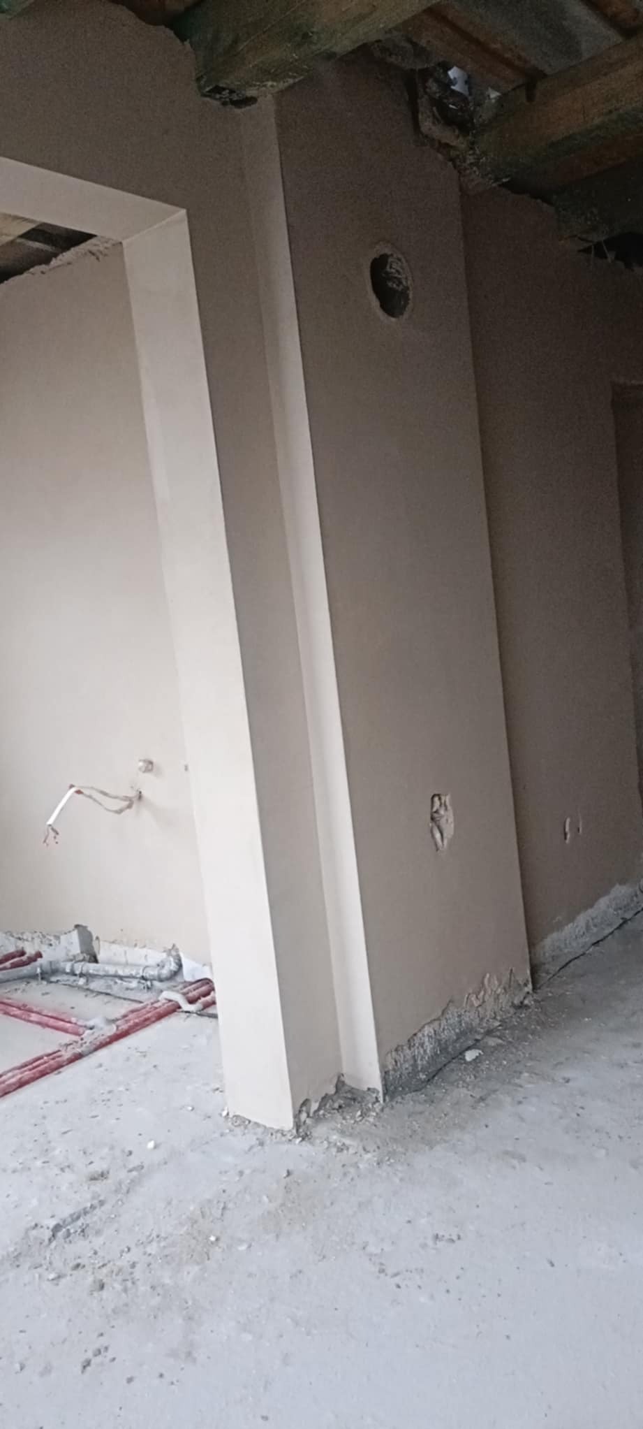 Świeżo otynkowane ściany w surowym wnętrzu, widoczne wystające kable elektryczne i rury instalacji wodnej, sufit z drewnianymi belkami.