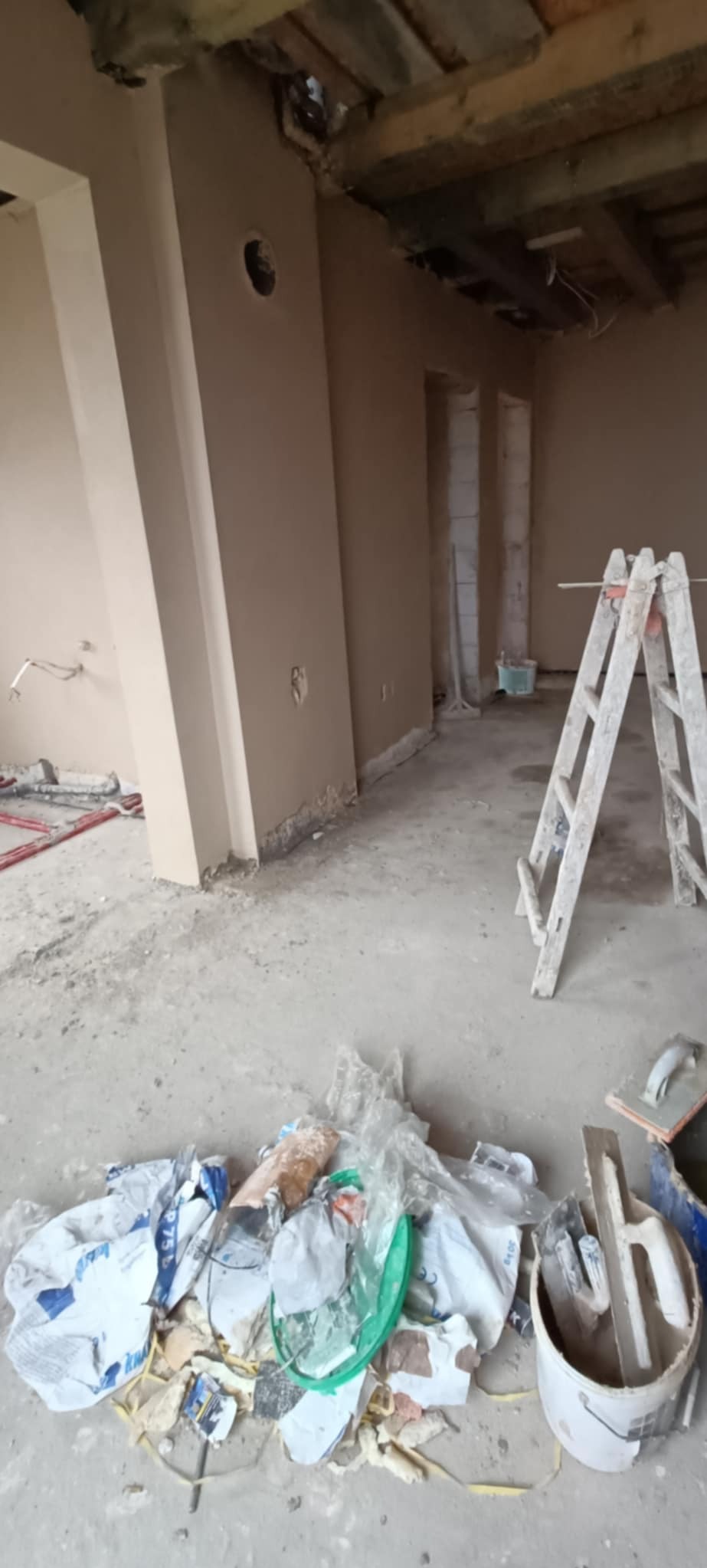 Wnętrze w trakcie prac wykończeniowych: świeżo otynkowane ściany, drewniany strop, drabina oparta o ścianę, na podłodze resztki materiałów budowlanych i narzędzia.