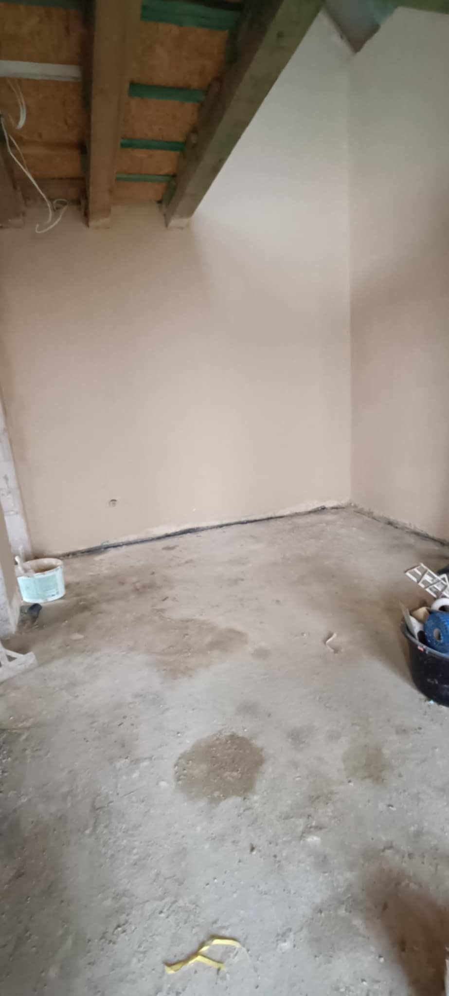 Pokój w trakcie prac wykończeniowych; świeżo otynkowane ściany w odcieniach beżu, surowy beton na podłodze z widocznymi plamami i zabrudzeniami, odsłonięta drewniana konstrukcja stropu.