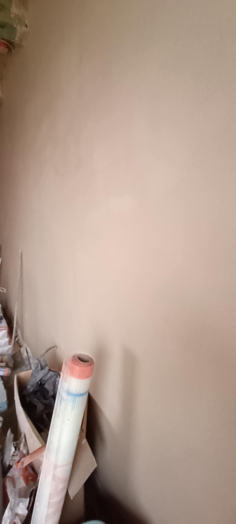 Gładka, beżowa ściana po nałożeniu tynku, z widocznym w rogu rolką siatki do zbrojenia i kartonem z narzędziami.