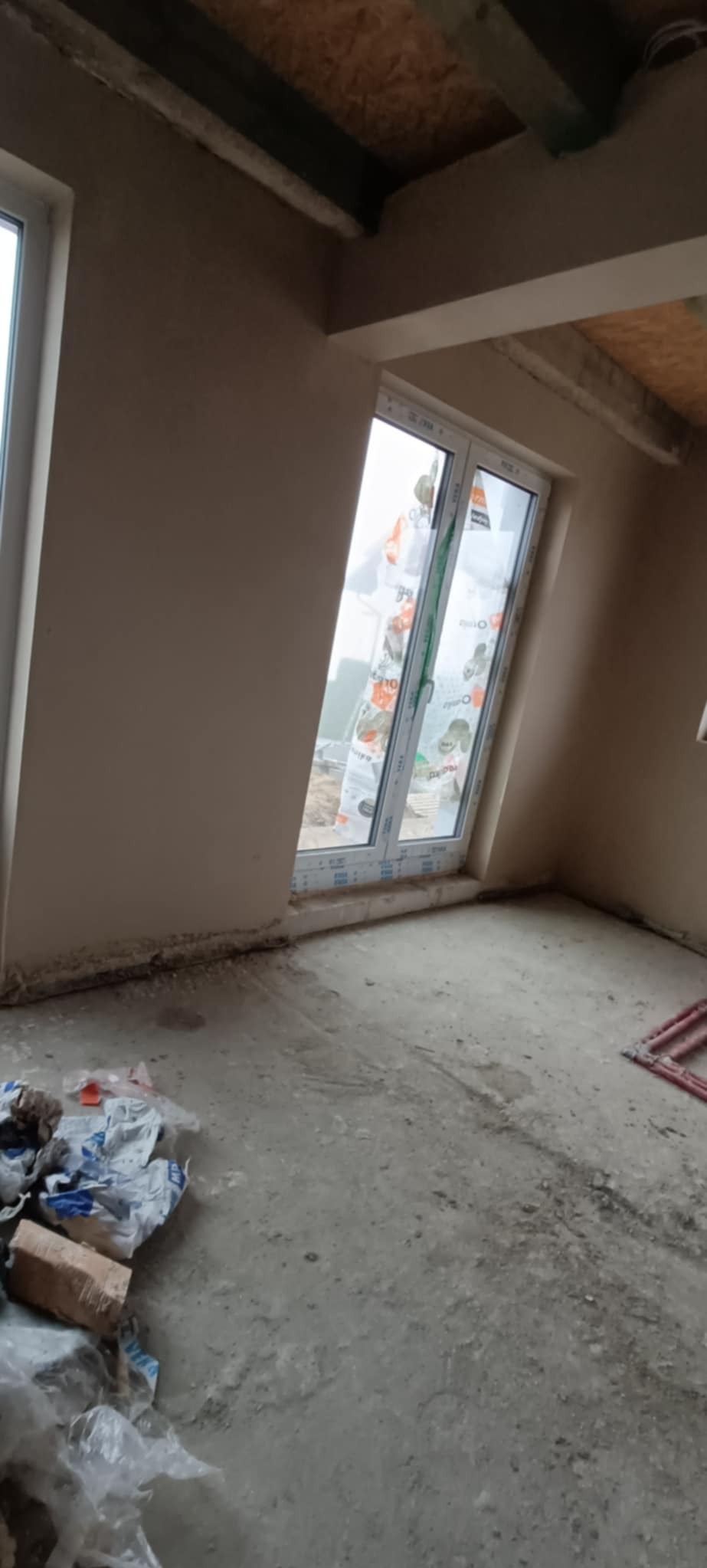 Surowe wnętrze po nałożeniu tynku, z widocznym oknem zabezpieczonym folią i nieuprzątniętym podłożem.