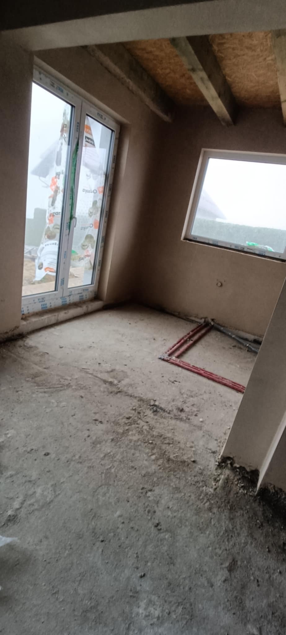 Surowe wnętrze pokoju w trakcie prac wykończeniowych, ściany pokryte świeżym tynkiem, widoczne okna PCV, instalacja grzewcza w podłodze i surowe drewniane belki stropowe.