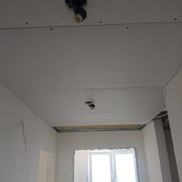 Sindbad - Montaż sufitu podwieszanego z płyt gipsowo-kartonowych, widoczne profile i instalacja elektryczna z oprawkami żarówek, ściany w trakcie szpachlowania, okno bez parapetu.