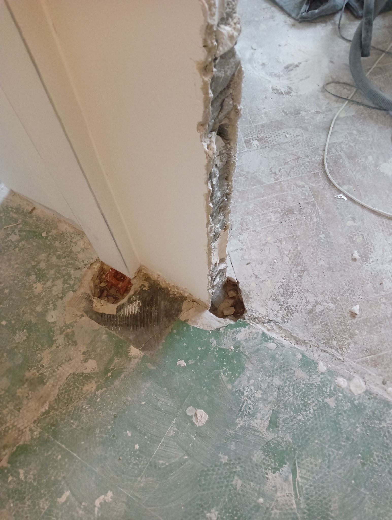 Demontaż starej futryny drzwiowej, widoczne fragmenty cegieł i resztki tynku na podłodze z zieloną farbą.