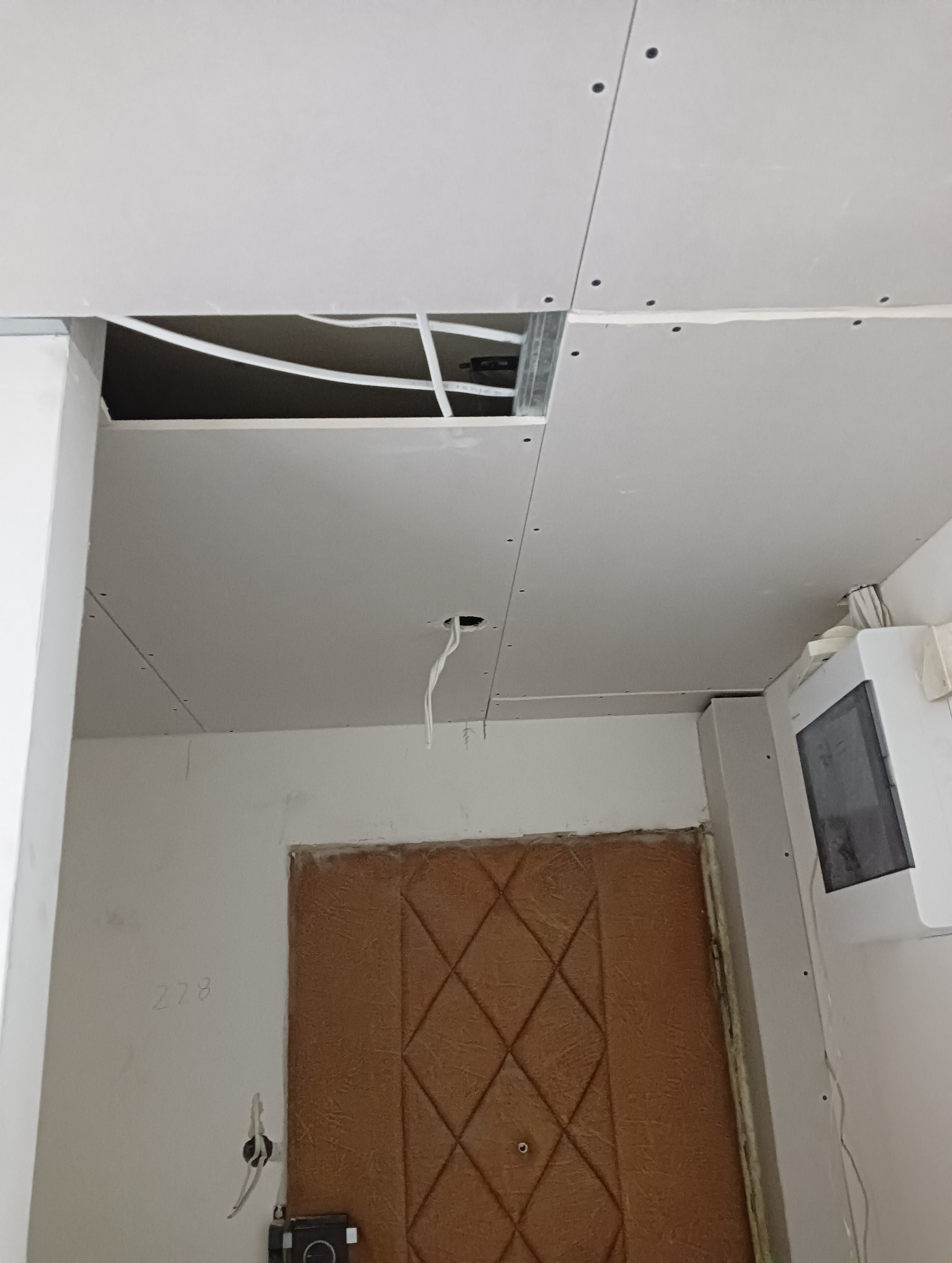 Montaż sufitu podwieszanego z płyt gipsowo-kartonowych, widoczne przewody elektryczne i otwór w płycie, brązowe drzwi wejściowe z geometrycznym wzorem w tle.