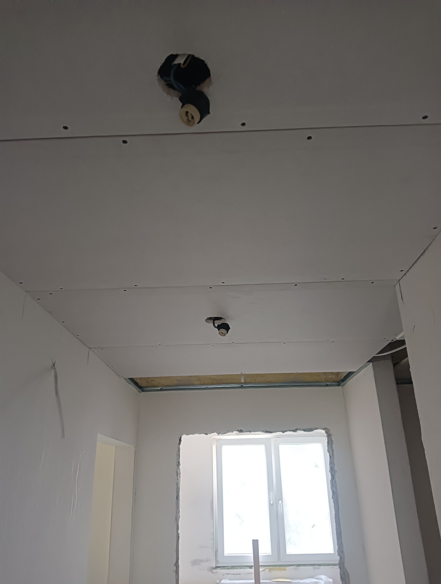 Montaż sufitu podwieszanego z płyt gipsowo-kartonowych, widoczne profile i instalacja elektryczna z oprawkami żarówek, ściany w trakcie szpachlowania, okno bez parapetu.