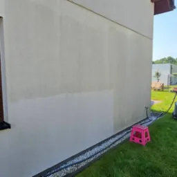 Fragment elewacji budynku z widoczną różnicą w czystości po częściowym umyciu, brązowa roleta okienna, myjka ciśnieniowa Karcher, różowy stołek na trawie.