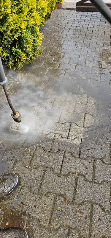 Czyszczenie kostki brukowej szarą myjką ciśnieniową z widocznym strumieniem wody i różnicą w kolorze między czystą i brudną kostką, obok zielony żywopłot.
