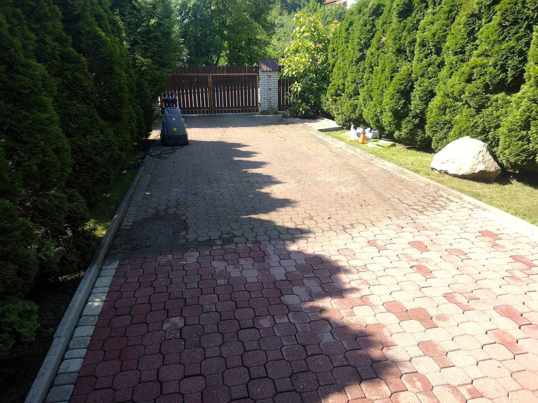 Częściowo wyczyszczona kostka brukowa na długim podjeździe, widoczna myjka ciśnieniowa, ciemne tuje po bokach i jasne plamy po czyszczeniu.
