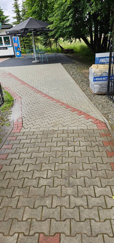 Częściowo oczyszczona, wilgotna kostka brukowa ułożona w zygzak, z widocznymi pozostałościami mchu w spoinach, prowadząca do kiosku z parasolem.