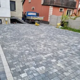 Ułożona szara kostka brukowa z jasnymi pasami, widoczny samochód dostawczy firmy brukarskiej i zagęszczarka w tle, narzędzia brukarskie przy krawędzi.