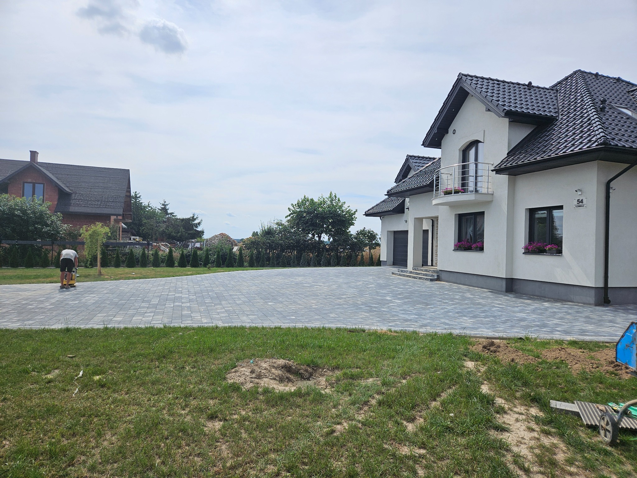 Nowoczesny dom jednorodzinny z szarą kostką brukową na podjeździe, zagęszczarką w użyciu i zielonym trawnikiem z fragmentami ziemi.