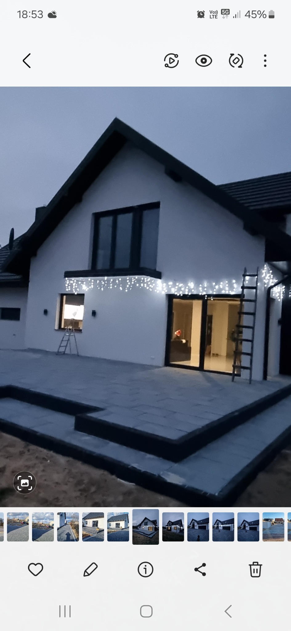 Nowoczesny dom z białymi ścianami i czarnym dachem, oświetlony girlandami świetlnymi, z tarasem wyłożonym szarą kostką, schodami i drabina oparta o ścianę.