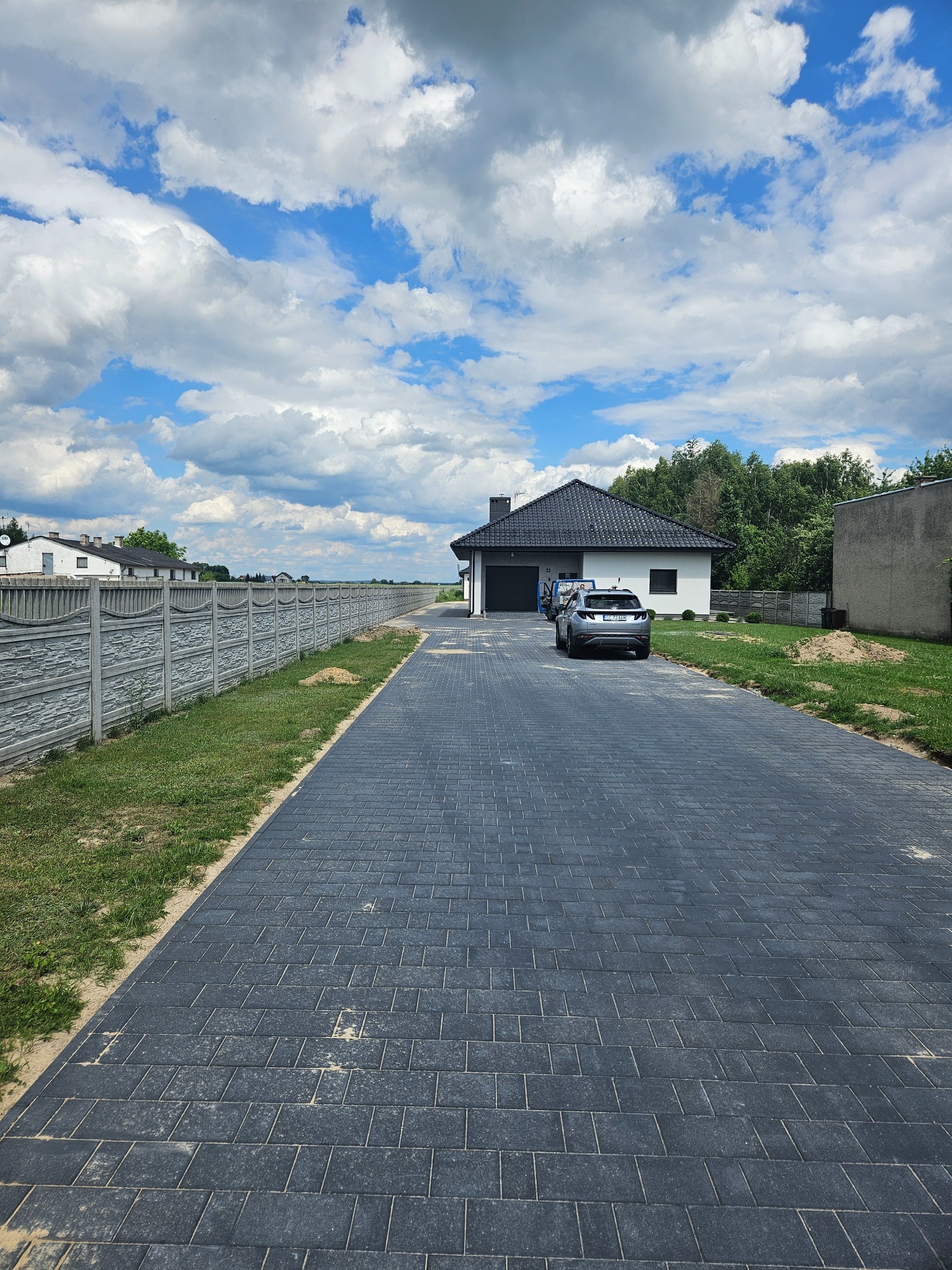Długa, ciemnoszara droga z kostki brukowej prowadząca do nowoczesnego domu z garażem, z betonowym ogrodzeniem po lewej stronie, samochód zaparkowany przed domem, błękitne niebo z obłokami w tle.