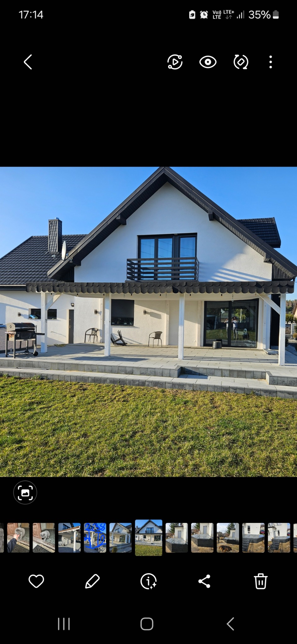 Nowoczesny dom z białymi ścianami, czarnym dachem i balkonem, otoczony zielonym trawnikiem i podjazdem z kostki brukowej.