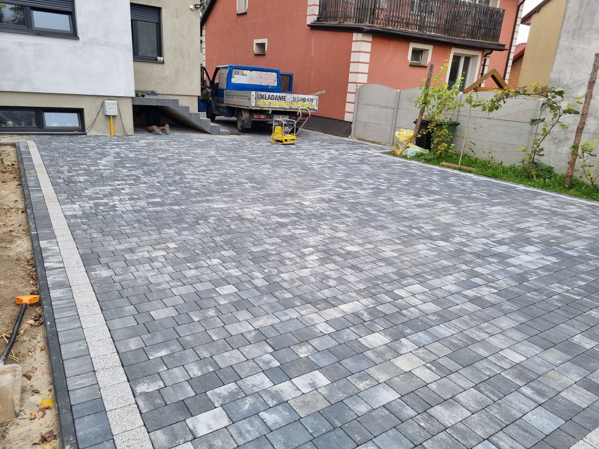 Ułożona szara kostka brukowa z jasnymi pasami, widoczny samochód dostawczy firmy brukarskiej i zagęszczarka w tle, narzędzia brukarskie przy krawędzi.